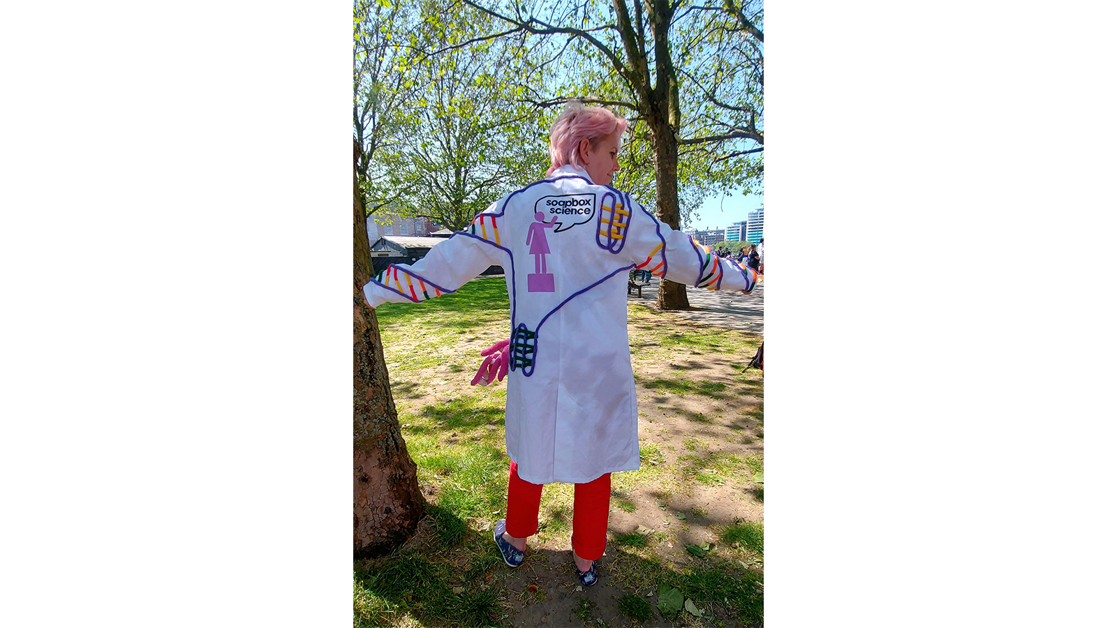 Zoe in a white coat with chemistry markings drawn on it, standing with back to us in sunshine
