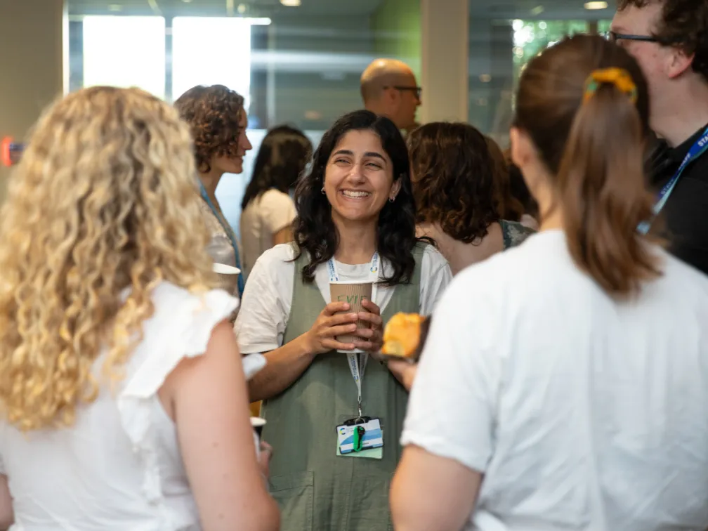 Candidate journey (image 2) A smiling woman with dark hair, holds a coffee cup while laughing and talking with two colleagues at a busy networking event.
