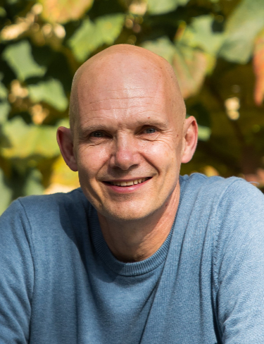 Iain McCulloch smiling warmly, wearing a long-sleeved, blue crew-neck sweater in an outdoor setting filled with green and yellowish leaves.