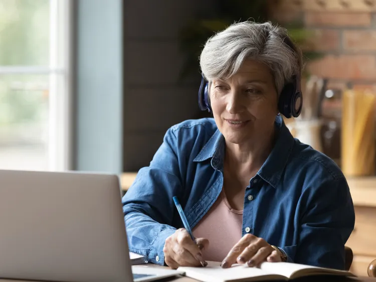 A woman with headphones writing on a notebook looking at laptop in front of her
