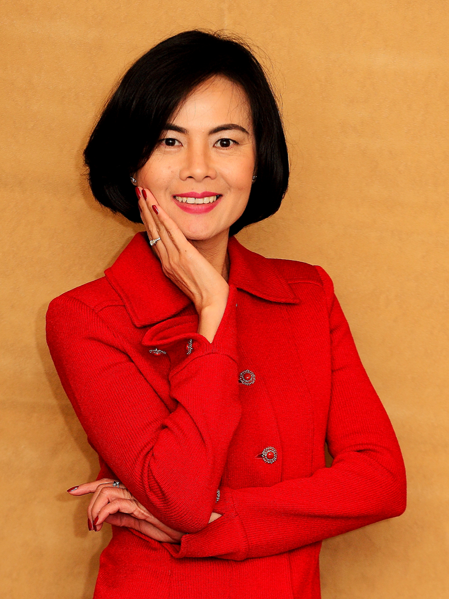 A studio portrait of Thuc-Quyen Nguyen smiling warmly, wearing a red coat, against a solid textured gold-tan background.