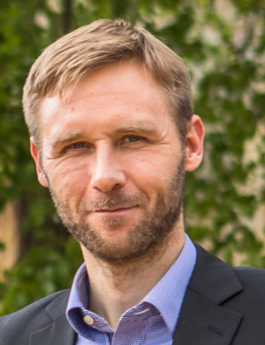Professor Tuomas Knowles smiling, wearing a dark suit jacket over a light blue button-down shirt in front of a green, leafy outdoor background.