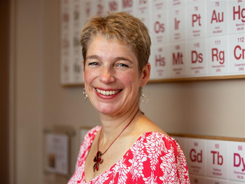 a woman smiling in front of a wall of periodic table