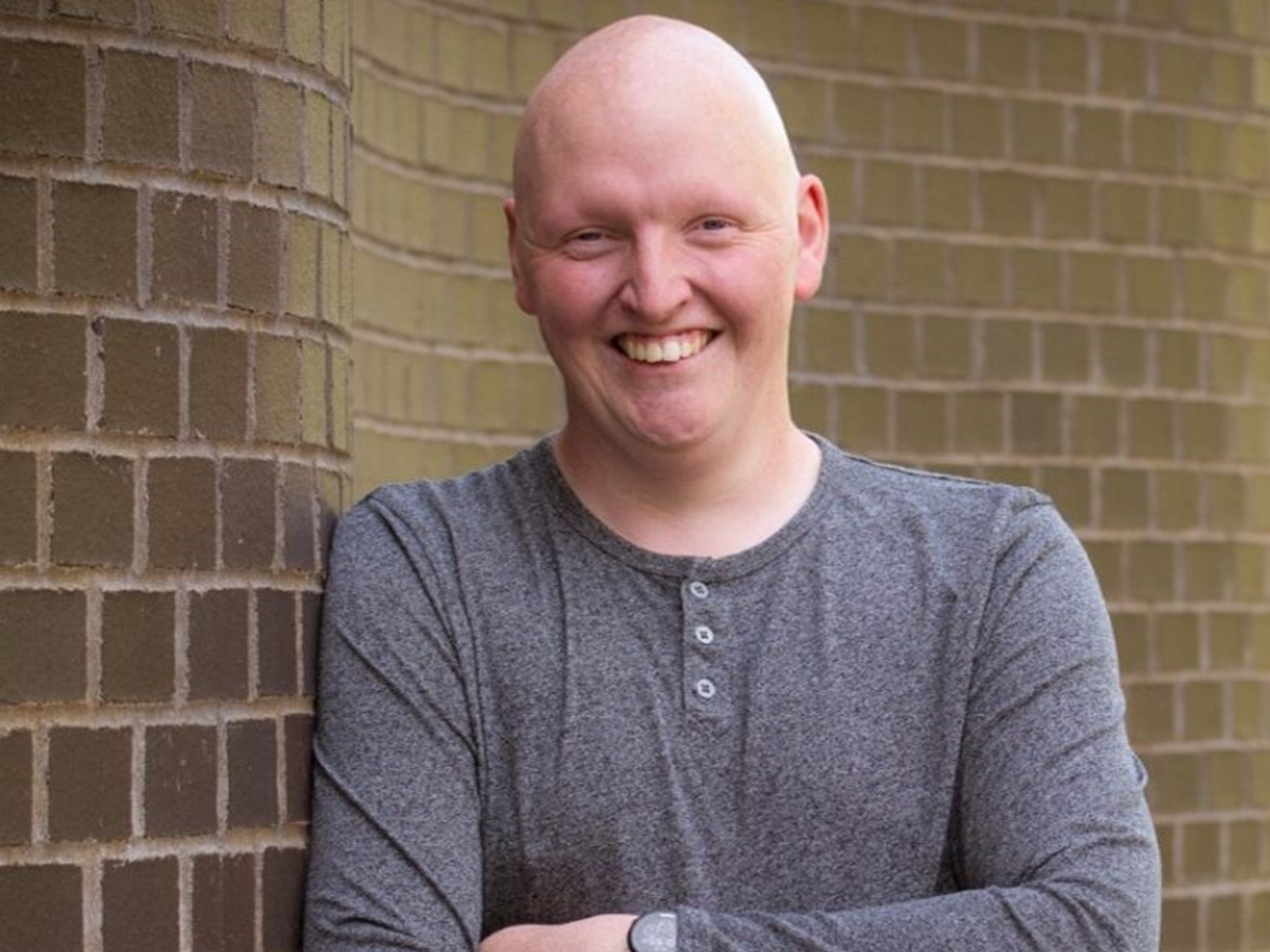 Dr Jack Woolley leaning against a brick wall outside, wearing grey jumper and smiling at camera