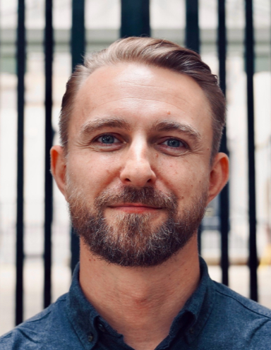 A headshot of Sam Cooper, smiling gently, wearing a dark blue button-down shirt and stands in front of a vertical black and white bar.