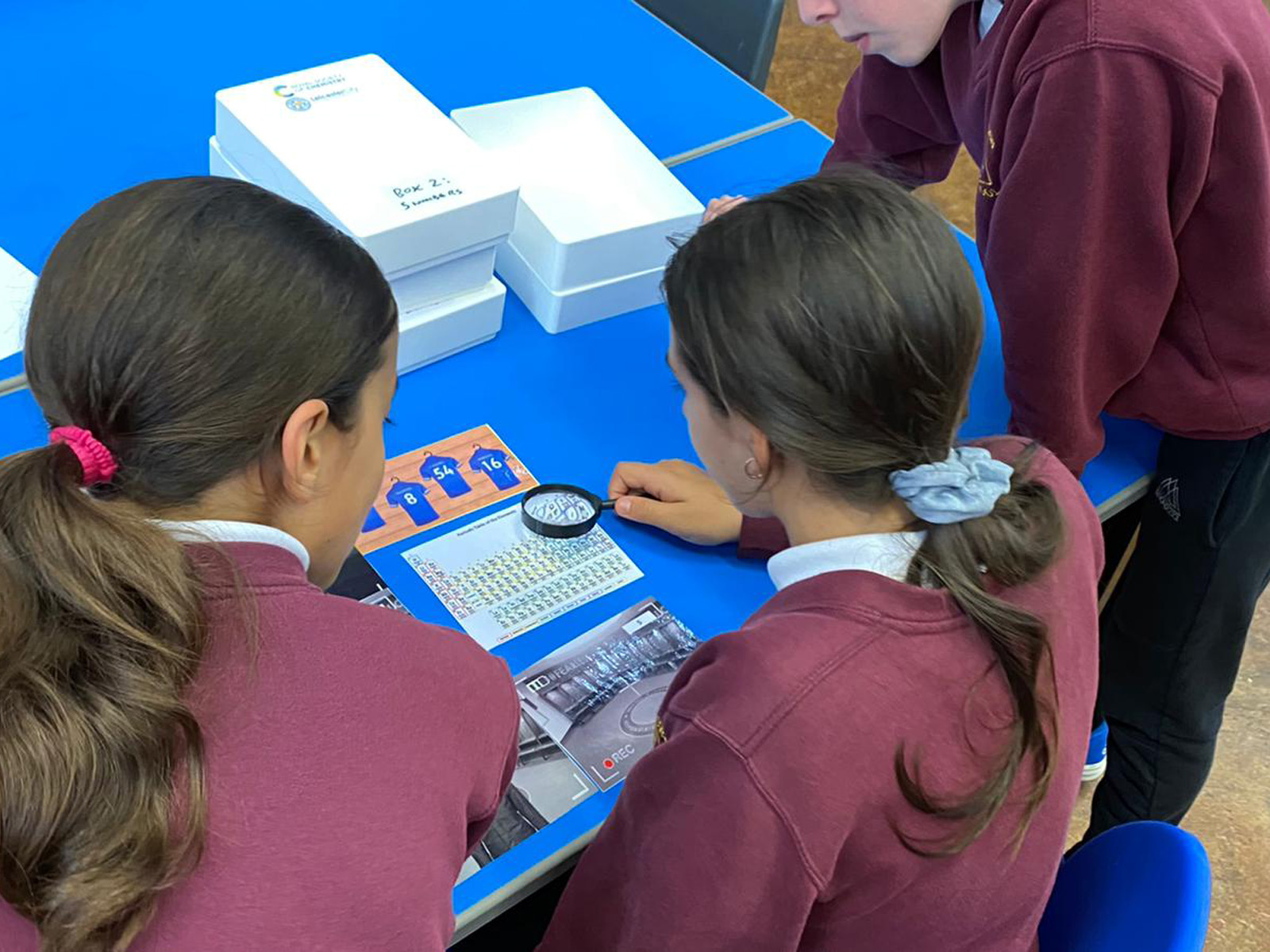 Schoolchildren use a magnifying glass to study a small copy of the periodic table