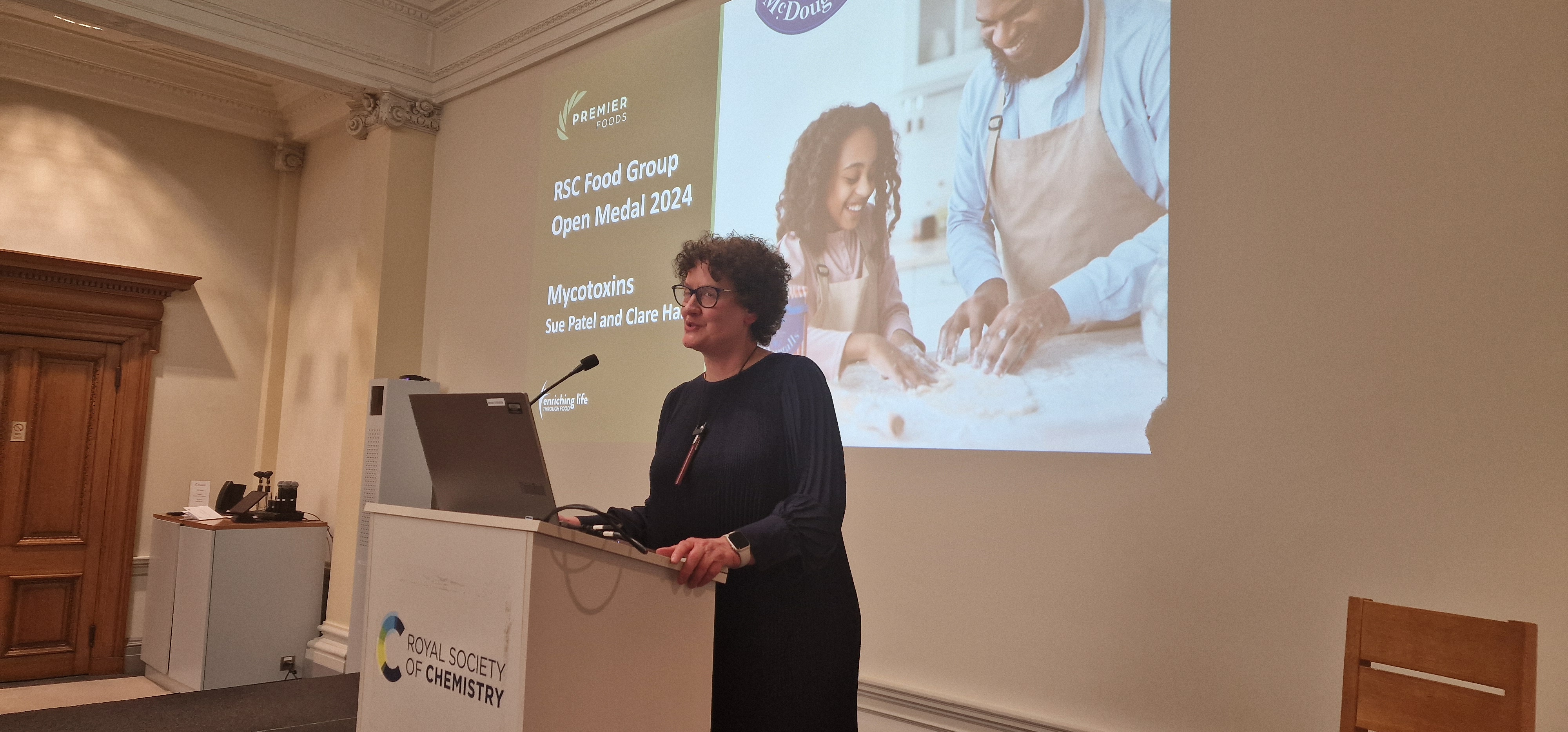 Dr Clare Hazel, of Premier Foods, stands behind a lectern with a graphic behind on a big screen, showing a father and daughter baking, as she prepares to give a presentation on mycotoxins