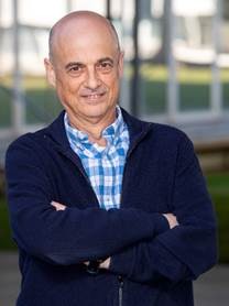 Antoni Llobet smiling, standing with his arms crossed, wearing a blue zip-up sweater over a blue and white checkered button-down shirt.