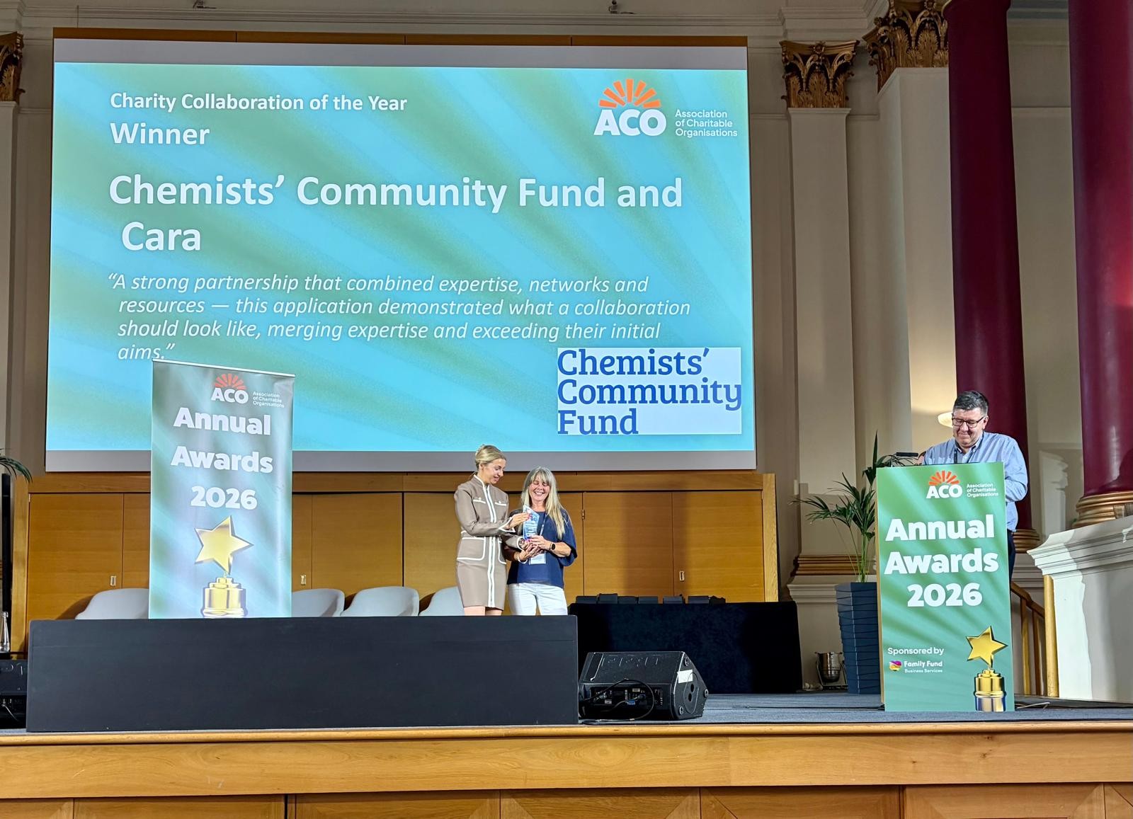 Chemists' Community Fund Senior Caseworker Dawn McGahan (centre) accepts the 2026 ACO Charity Collaboration of the Year award from a woman to her left as a man stands behind a lectern to the right.