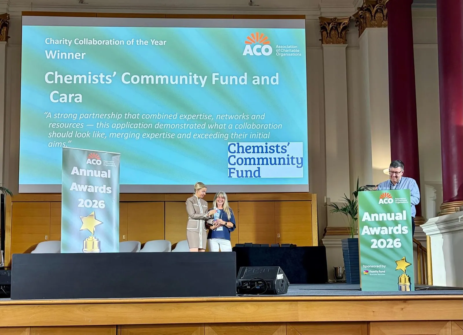 ACO CCF & Cara Award Presentation Chemists' Community Fund Senior Caseworker Dawn McGahan (centre) accepts the 2026 ACO Charity Collaboration of the Year award from a woman to her left as a man stands behind a lectern to the right.