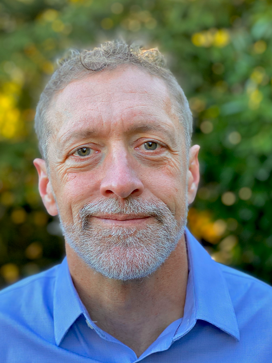Rob Britton smiling to camera, wearing bright blue shirt, he has grey beard, moustache and hair.