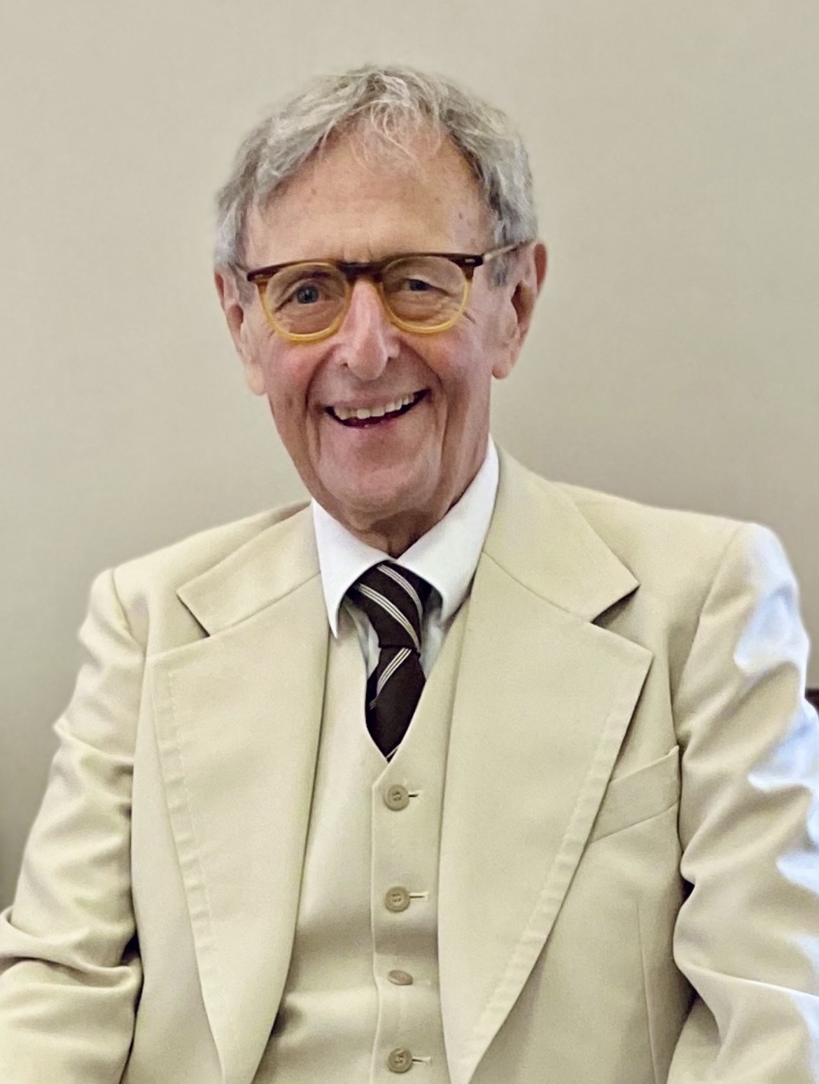 Kenneth Wallace Smalldon smiles widely to camera in 3 piece cream suit and tie and wearing glasses