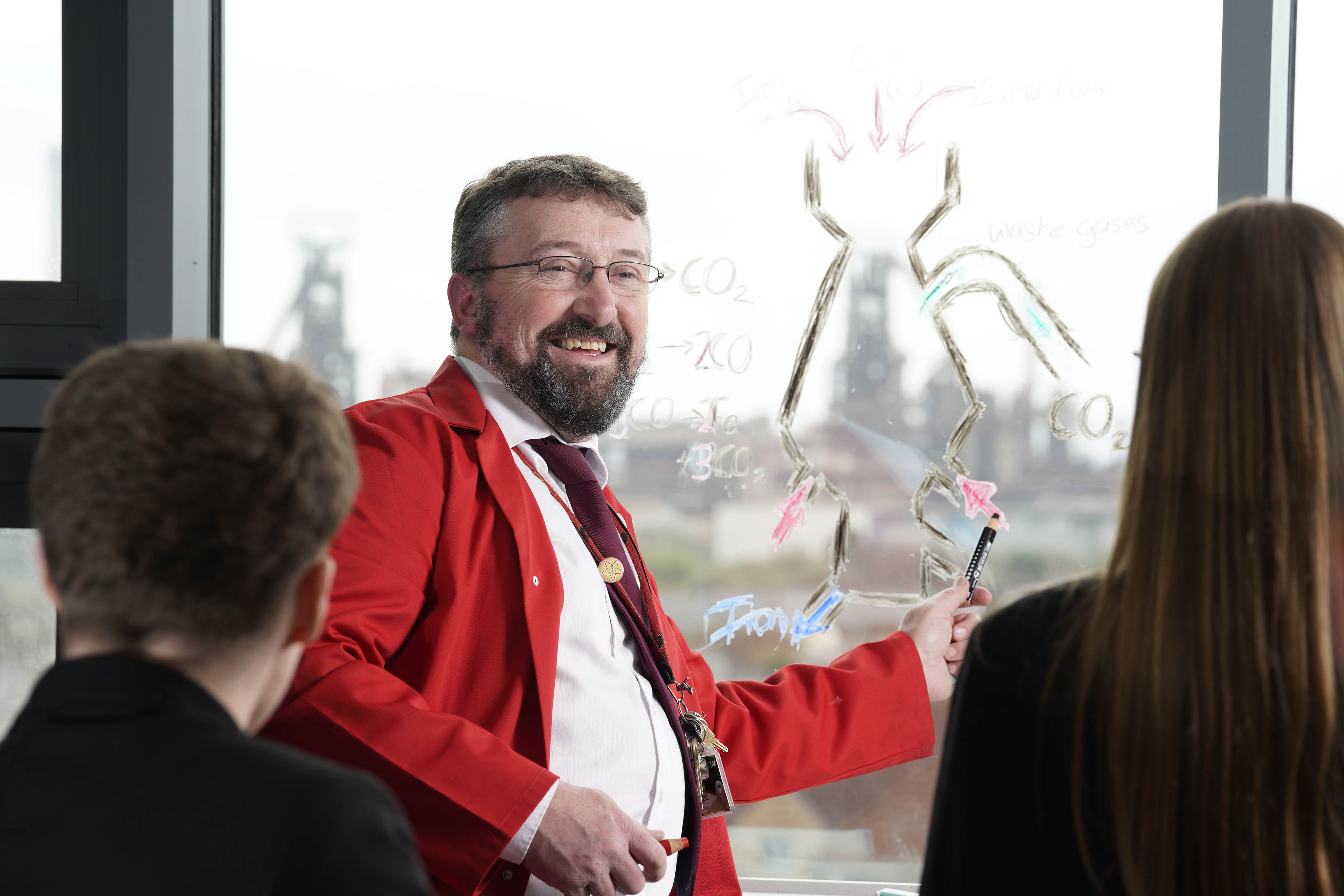 This image shows a teacher in a red jacket explaining a diagram drawn on a glass pane to others.