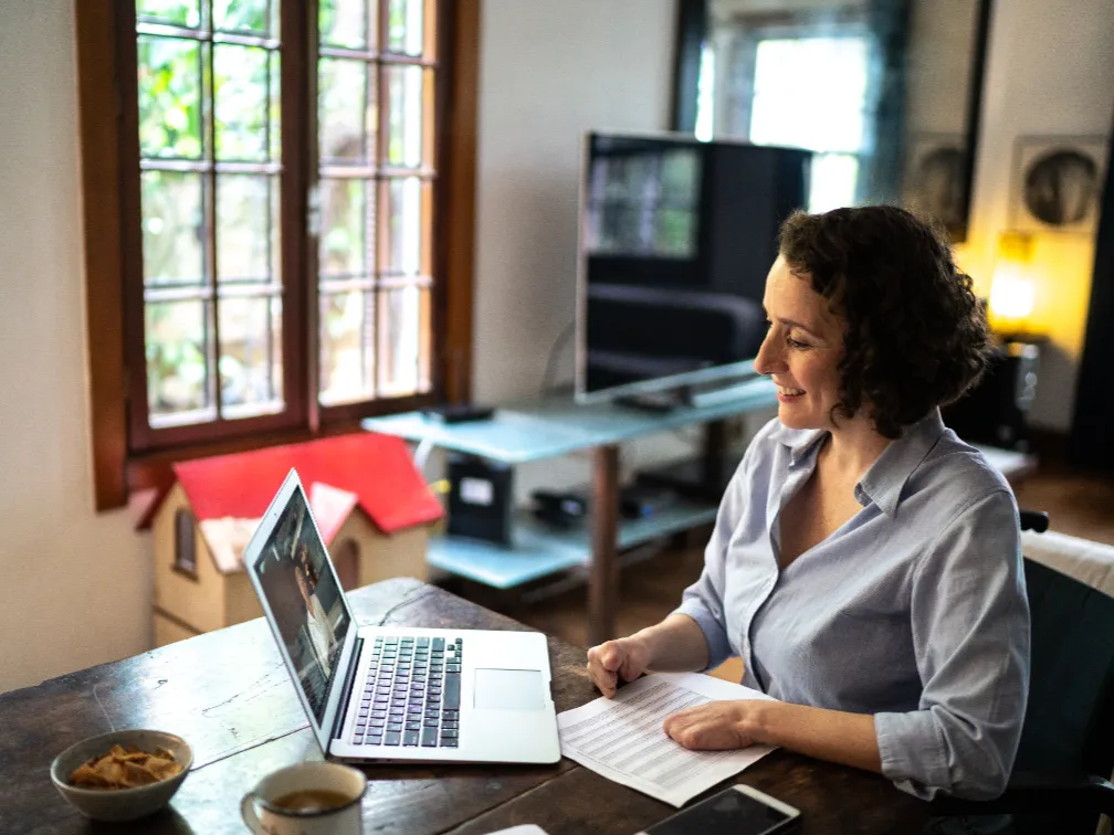 Candidate journey (image 3) A woman with curly brown hair sits at a rustic wooden table, smiling while looking at her open laptop during a video call.