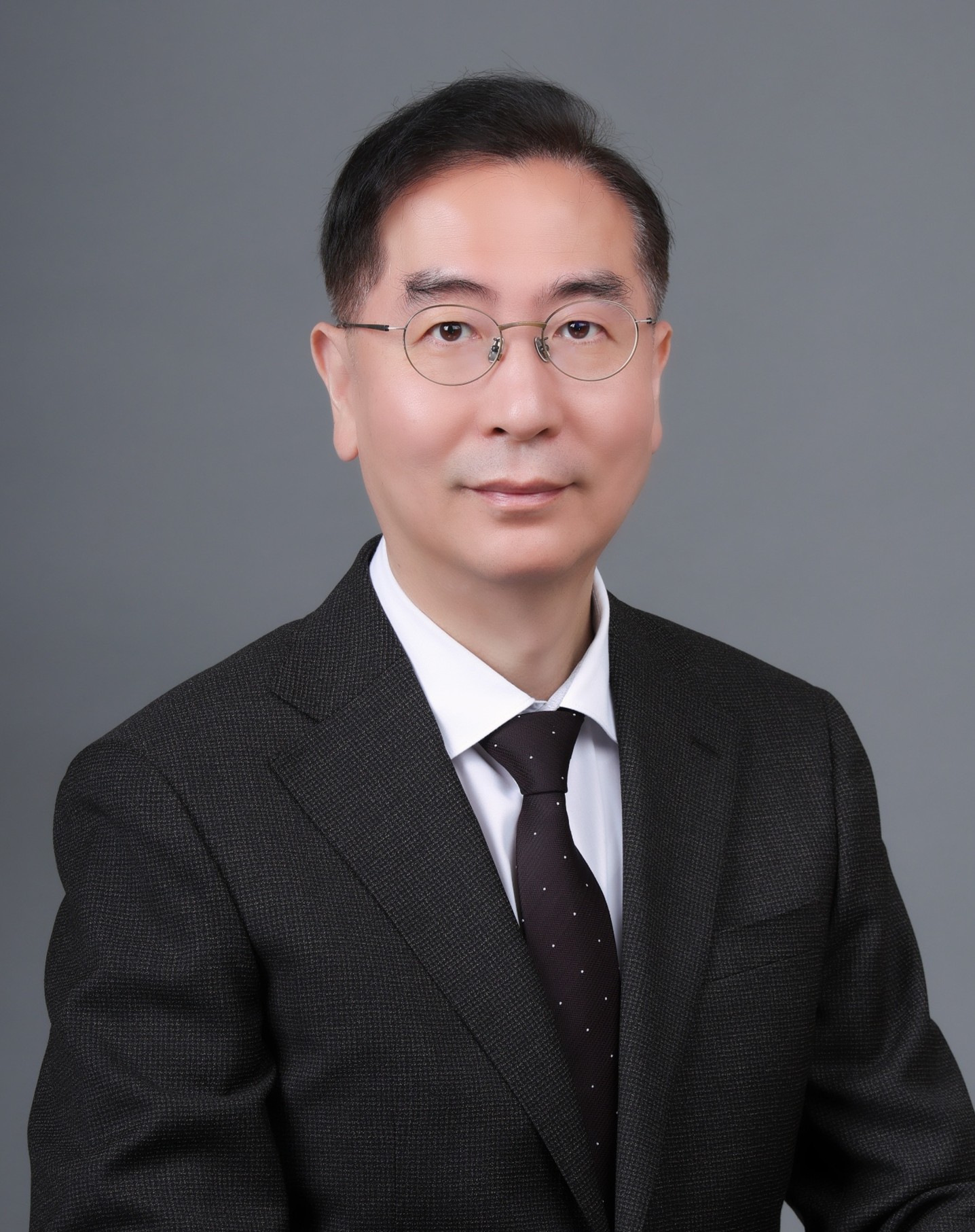 Dong-Chan Oh smiling faintly and dressed formally in a dark suit jacket, and tie against a gray background.