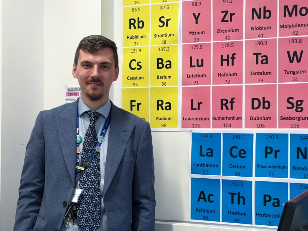 Matthew Gundry smiling to camera in suit and tie in front of wall with periodic table