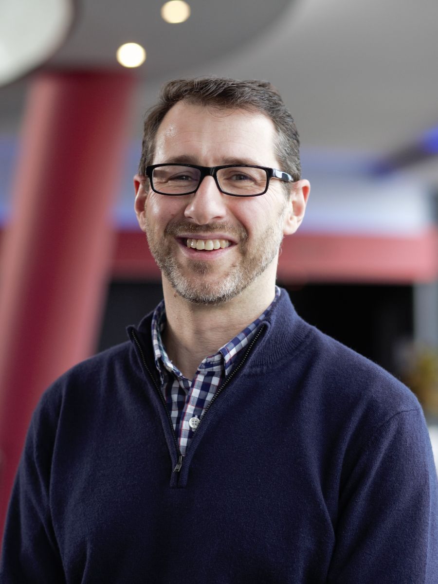 Nicholas Dunne smiling to camera wearing glasses, beard, check shirt