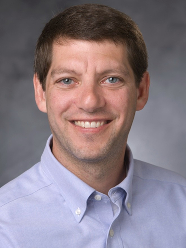 Headshot of Andrew B Nixon, smiling warmly while wearing a light blue button-down shirt against a neutral grey background.