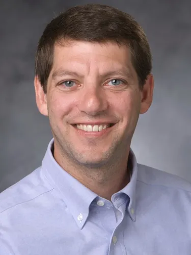 Andrew B Nixon Headshot of Andrew B Nixon, smiling warmly while wearing a light blue button-down shirt against a neutral grey background.