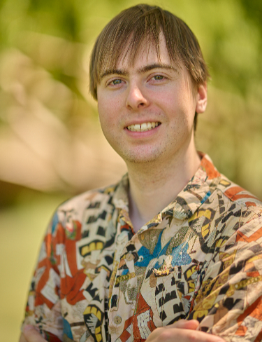 James Ward Taylor smiling, wearing a graphic-print button-down shirt against a softly blurred green background.