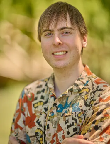 James Ward Taylor James Ward Taylor smiling, wearing a graphic-print button-down shirt against a softly blurred green background.