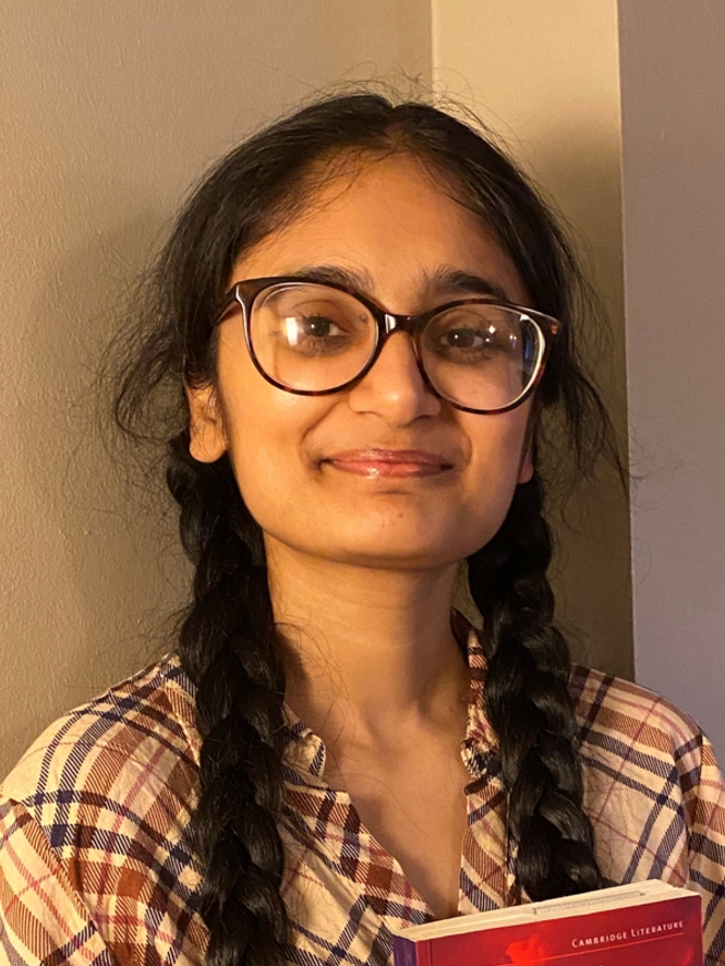 Manpreet Kaur smiling with braided hair wearing glasses and a plaid shirt while holding a book.