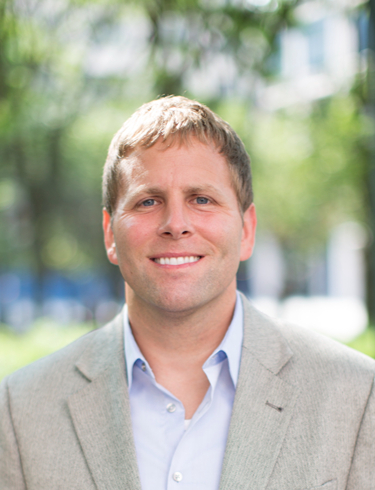 Ryan L Hartman smiling at the camera, wearing a light blue button-down shirt under a light gray textured blazer. 