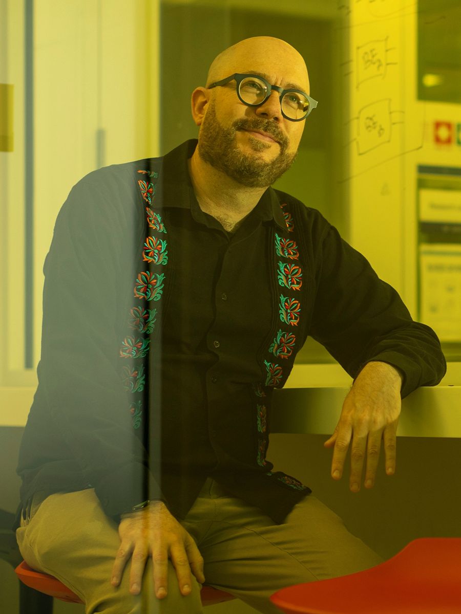 Alán Aspuru-Guzik sitting on stool leaning on shelf in lecture lab, wearing glasses and black shirt with coloured patterned squares