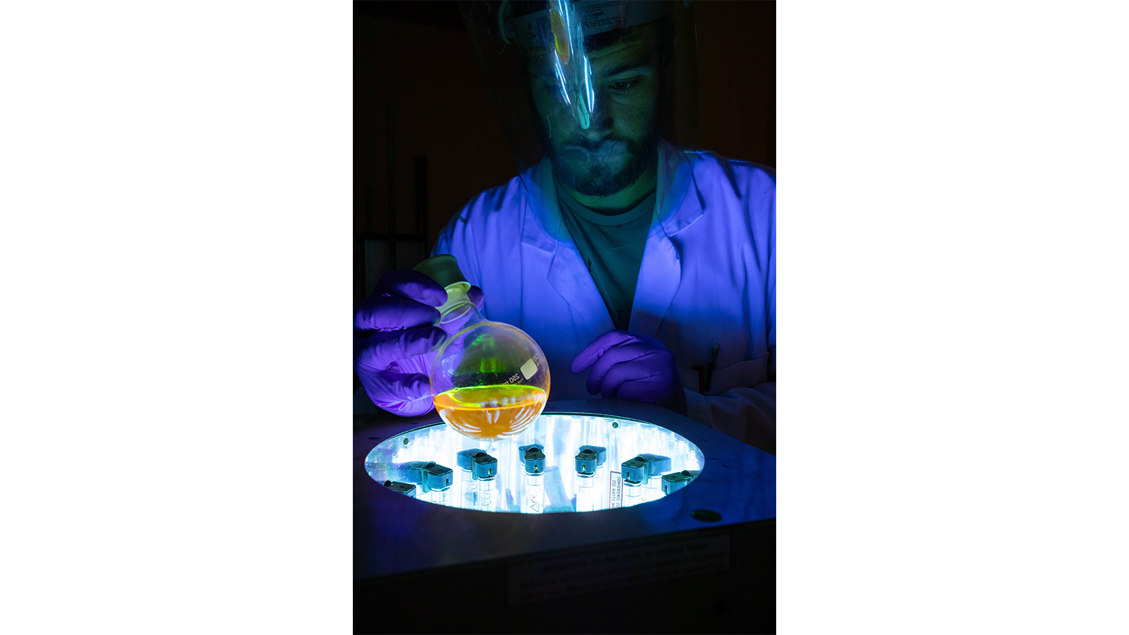 PISCO Team member in a white coat and gloves holding a flask with yellow liquid in blue light