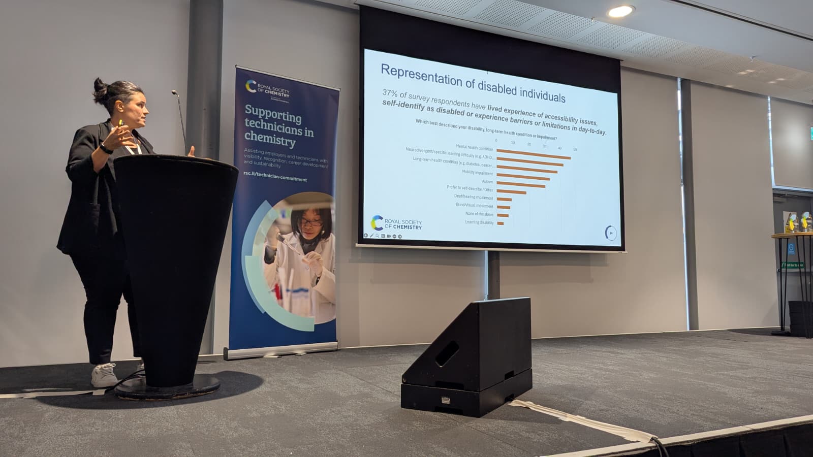 Dr Ilaria Meazzini, Senior Programme Manager for Inclusion & Diversity at the Royal Society of Chemistry, stands beside a podium in front of a screen