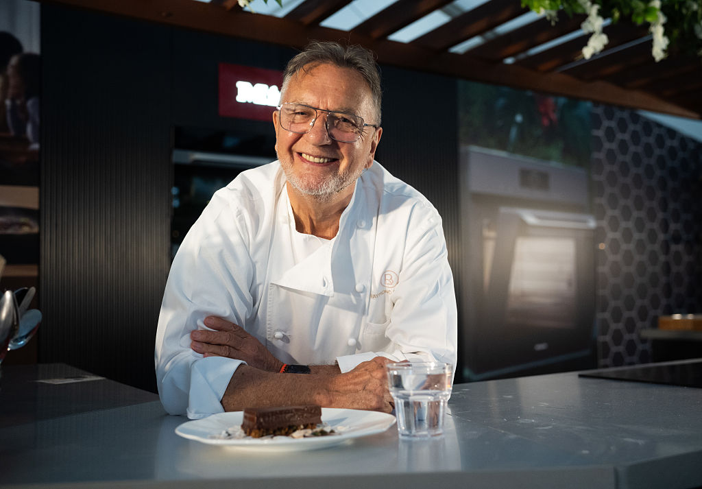 Raymond Blanc smiles and perches on a work surface in a big kitchen