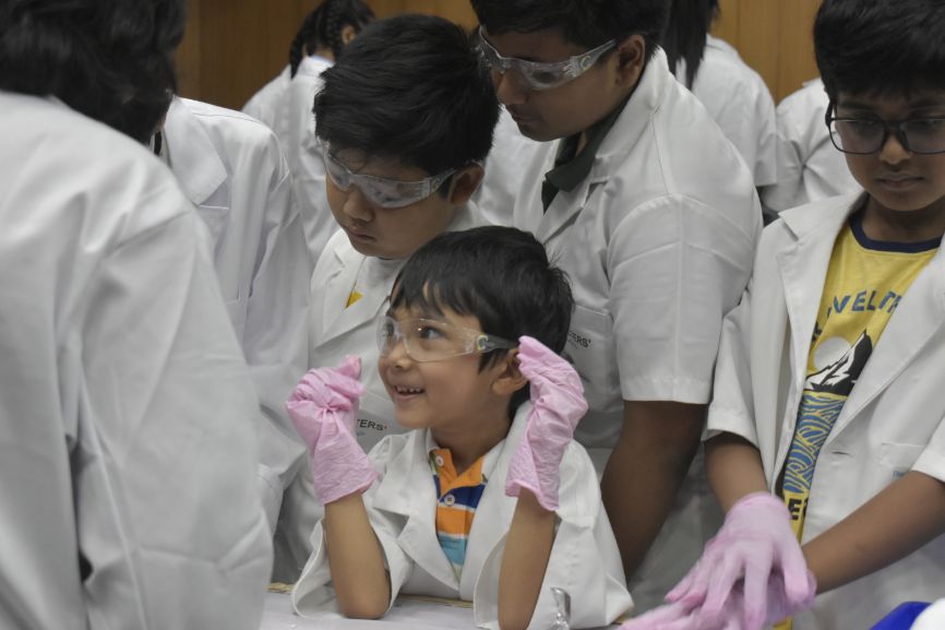 An excited young boy wearing safety goggles and gloves watches an experiment with other children