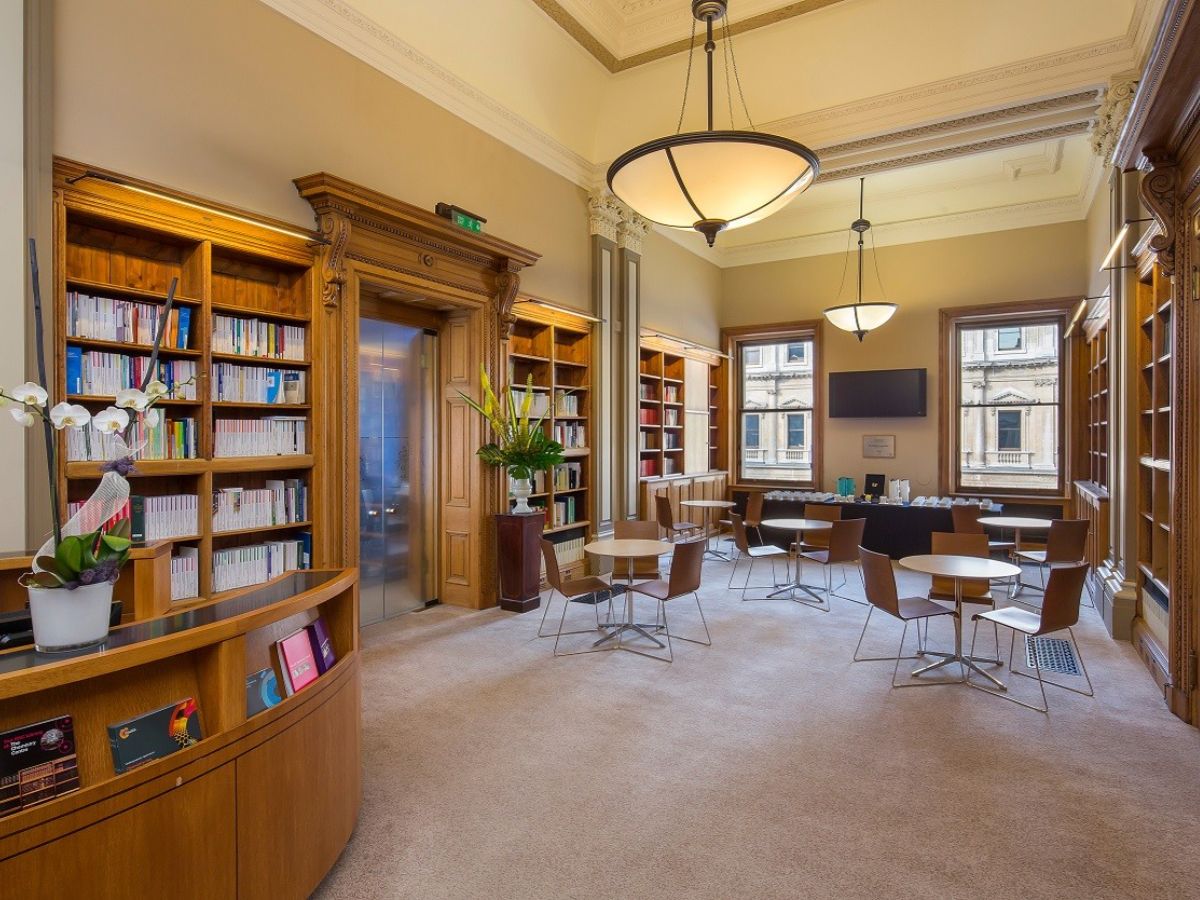 A library desk in an older classic style room with many book shelves and tables and chairs