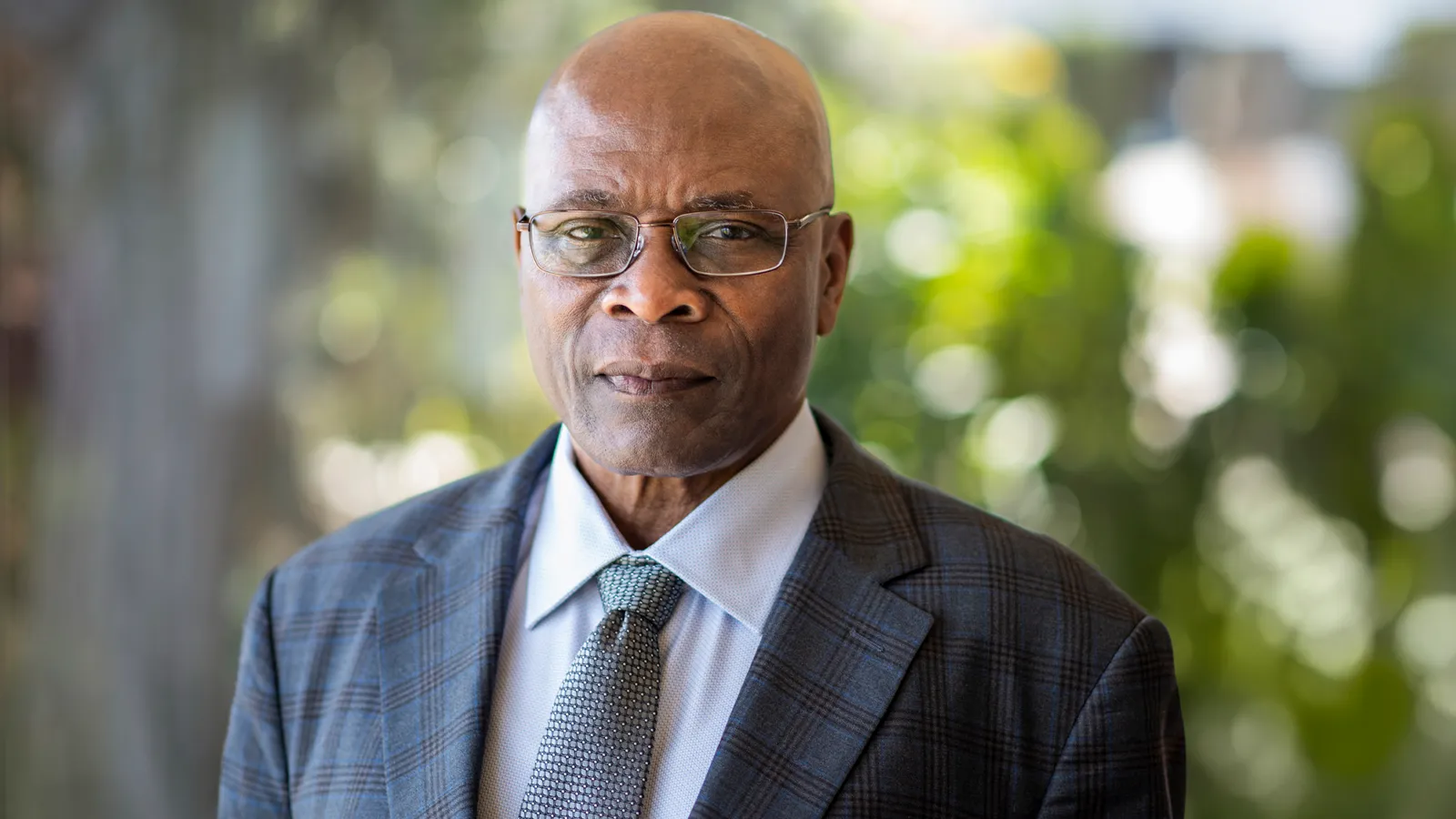 Professor Samson Jenekhe wearing a suit and tie and glasses against a blurred background of foliage