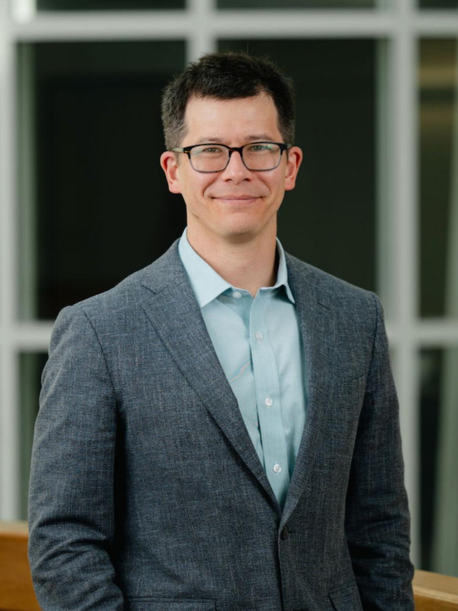 Keary in a grey suit and shirt smiling to camera wearing glasses