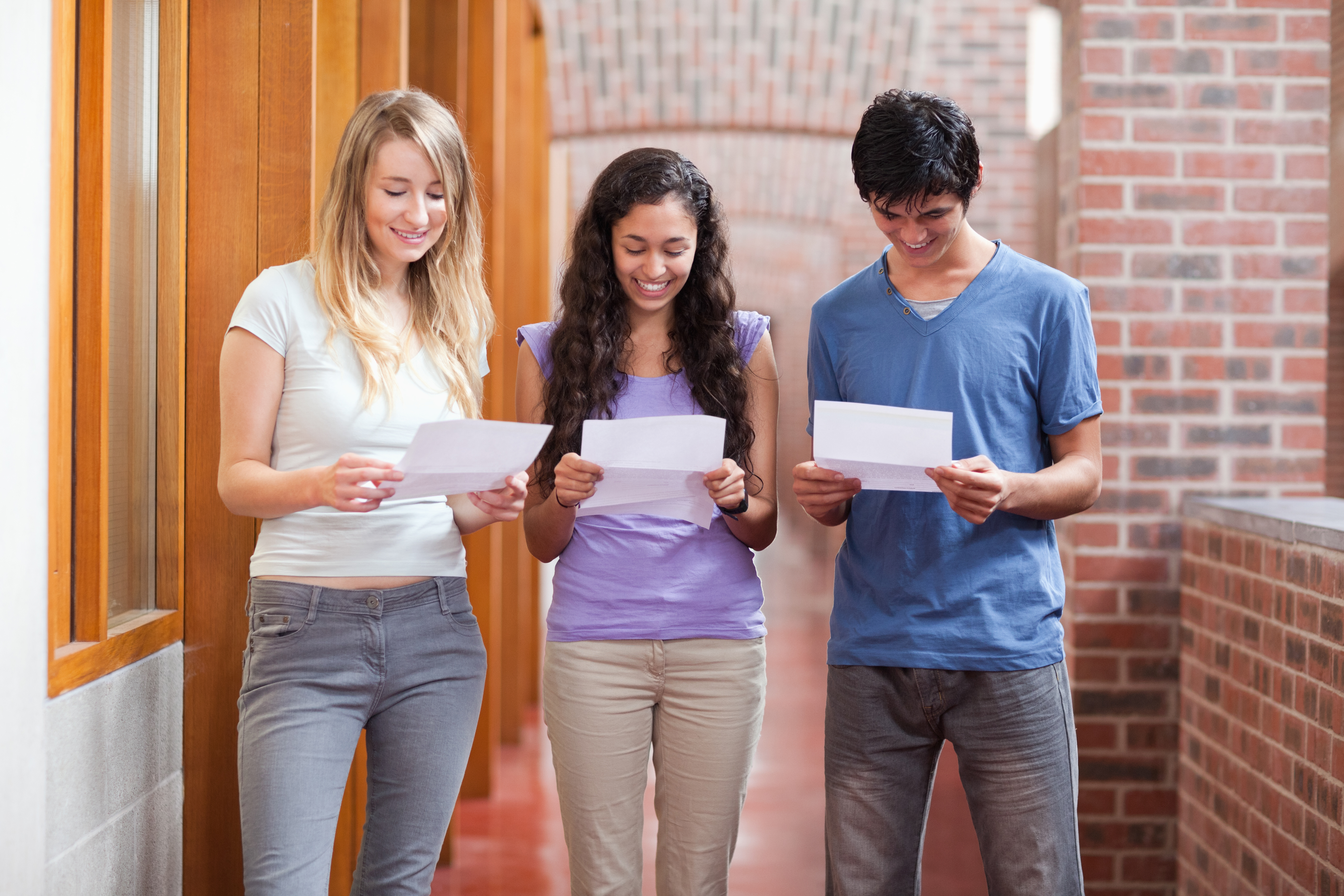 Three high school students, two female and one male, stand together looking down at pieces of paper with their exam results on them
