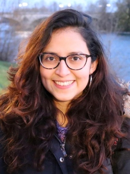 Andrea Carolina Ojeda-Porras smiling with long dark hair wearing glasses and a dark jacket outdoors.