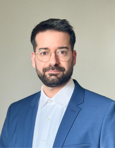 Professor Alexandros Sofias wearing round-rimmed eyeglasses, white button-down shirt, and a structured blue blazer against a neutral backdrop.