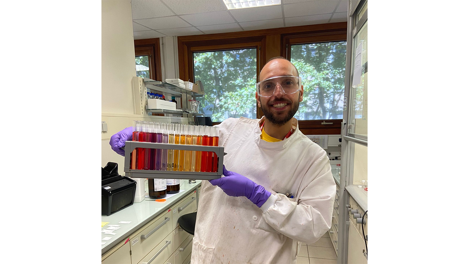 PISCO Team member in a lab coat and gloves holding a tray of test tubes containing coloured liquids