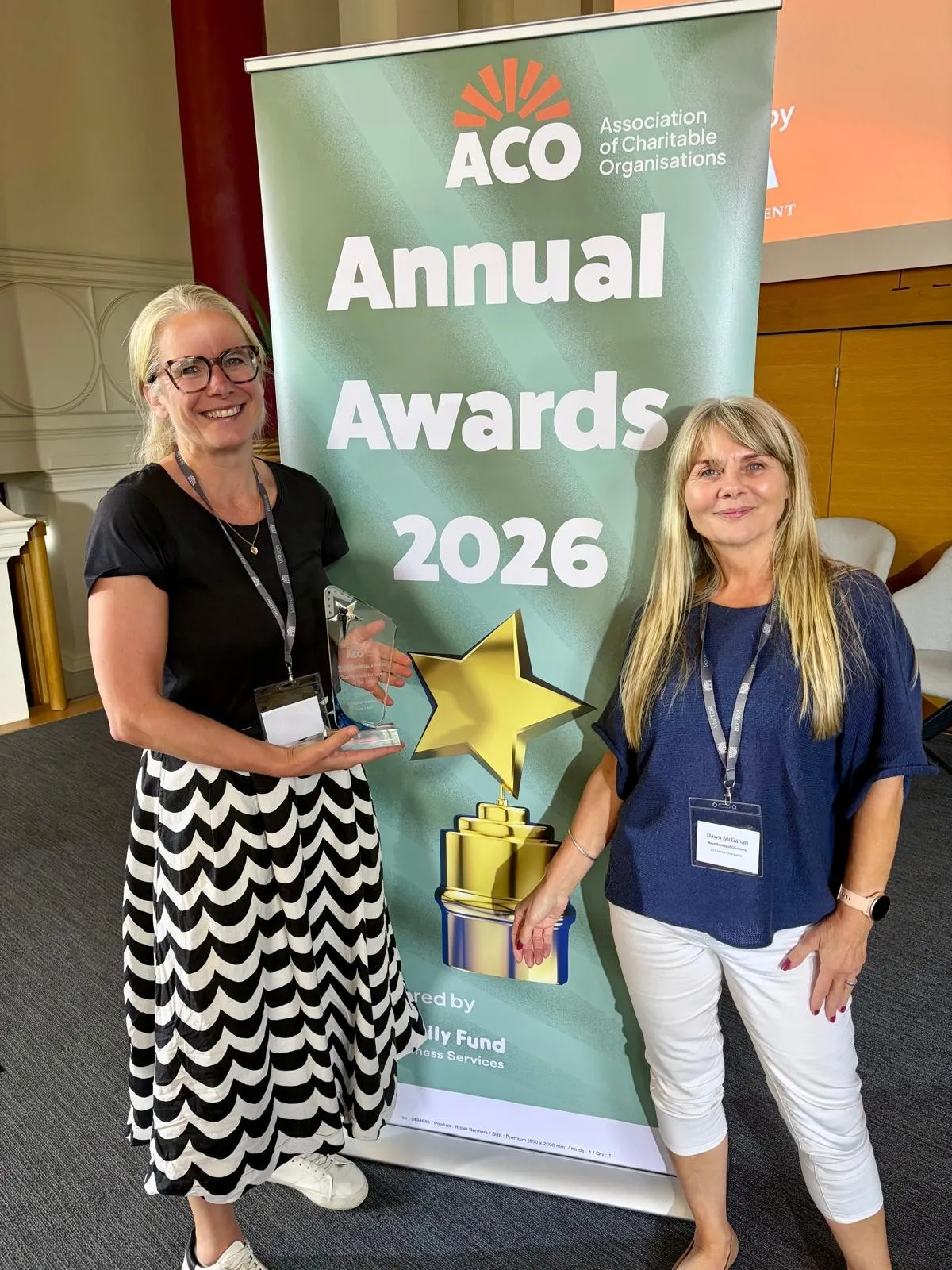 Maggie & Dawn ACO Award Maggie Houghton and Dawn McGahan standing next to a sign reading "ACO Annual Awards 2026" with Maggie holding a glass trophy