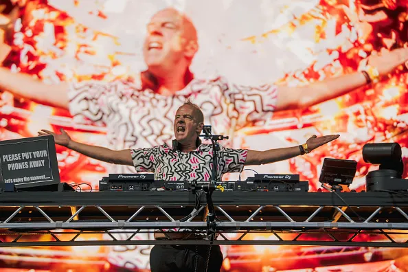 GettyImages-2287519035 - Fatboy Slim Fatboy Slim aka Norman Cook stands with his arms raised on a stage with a broadcast of himself on a huge screen behind him
