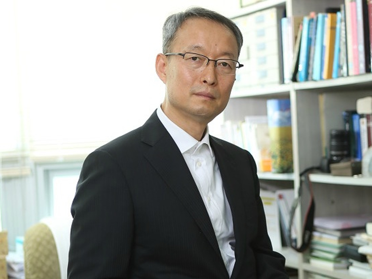 A medium close-up shot of Paik Ungyu, sitting in an office setting. He is wearing a dark suit jacket, a white collared shirt and glasses.