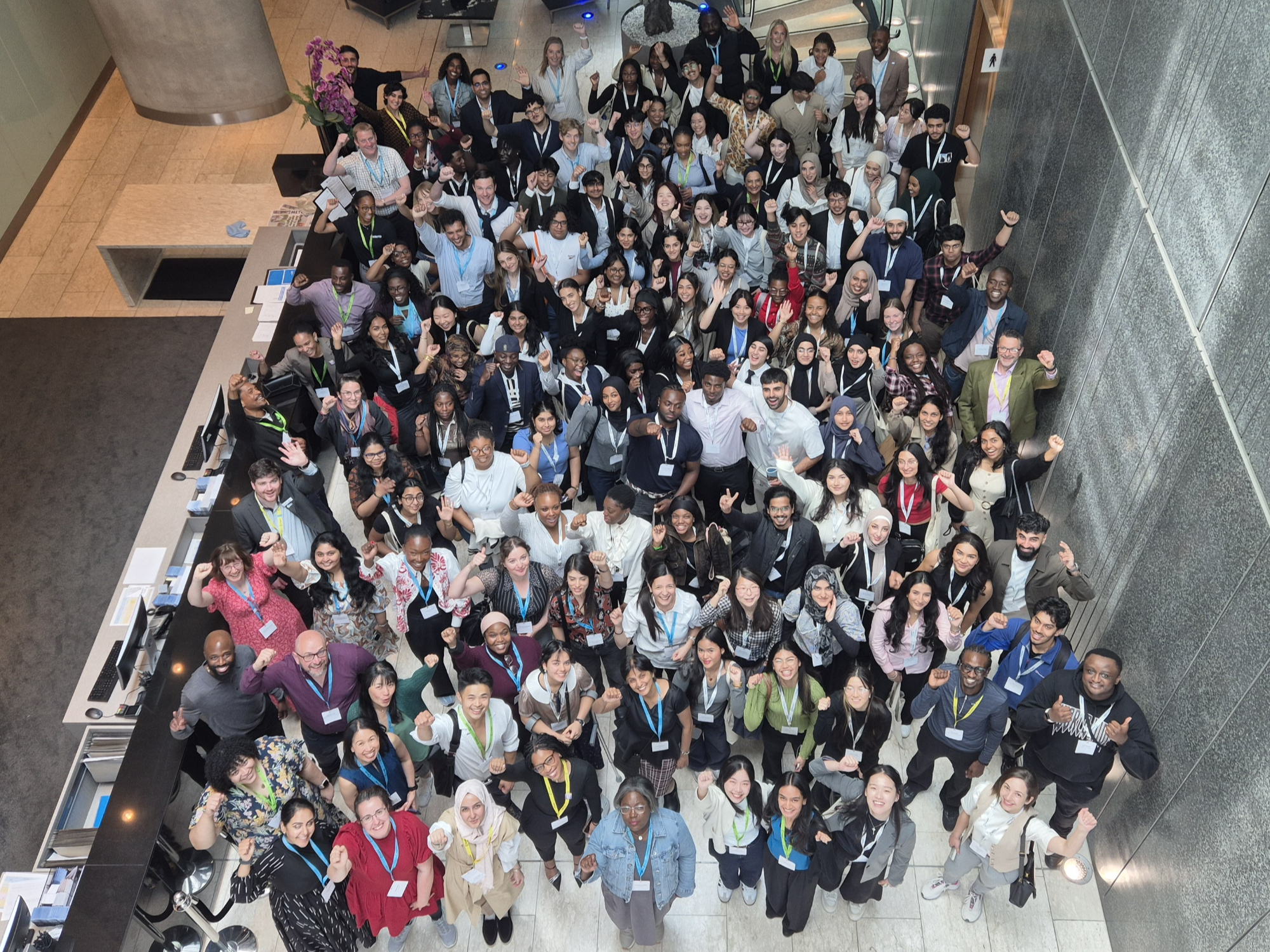 The students from the Broadening Horizons cohort grouped in a large hall waving and smiling, with photo taken from mezzanine looking down on them