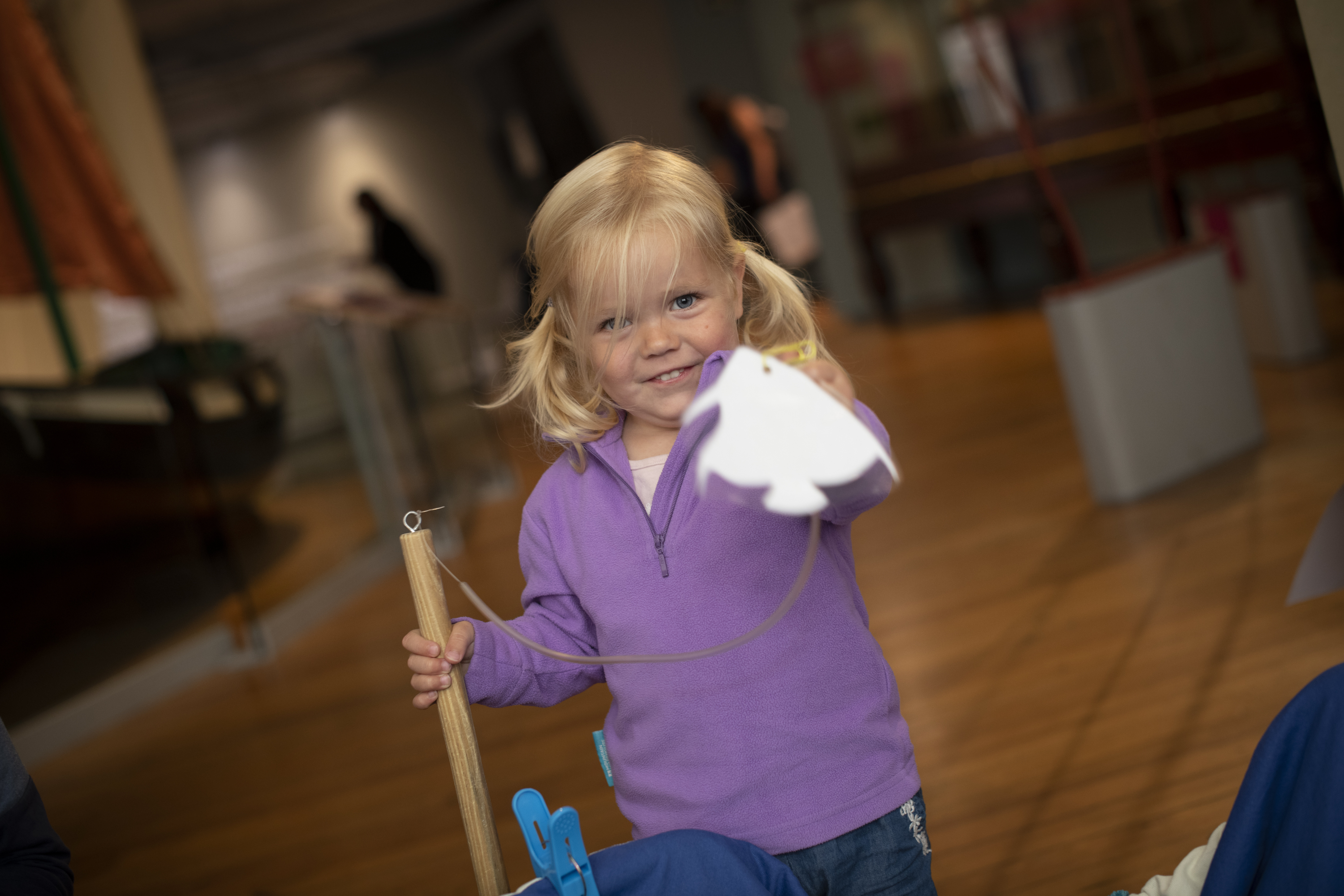 A little girl in a purple jumper points at the camera while holding something in her left hand and gripping a wooden post with her right.