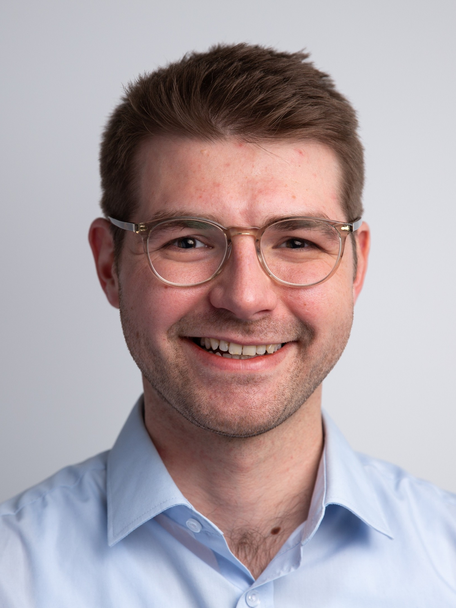 Edward Gardner smiling at the camera, wearing round, clear-framed glasses and a light blue shirt against a grey background.
