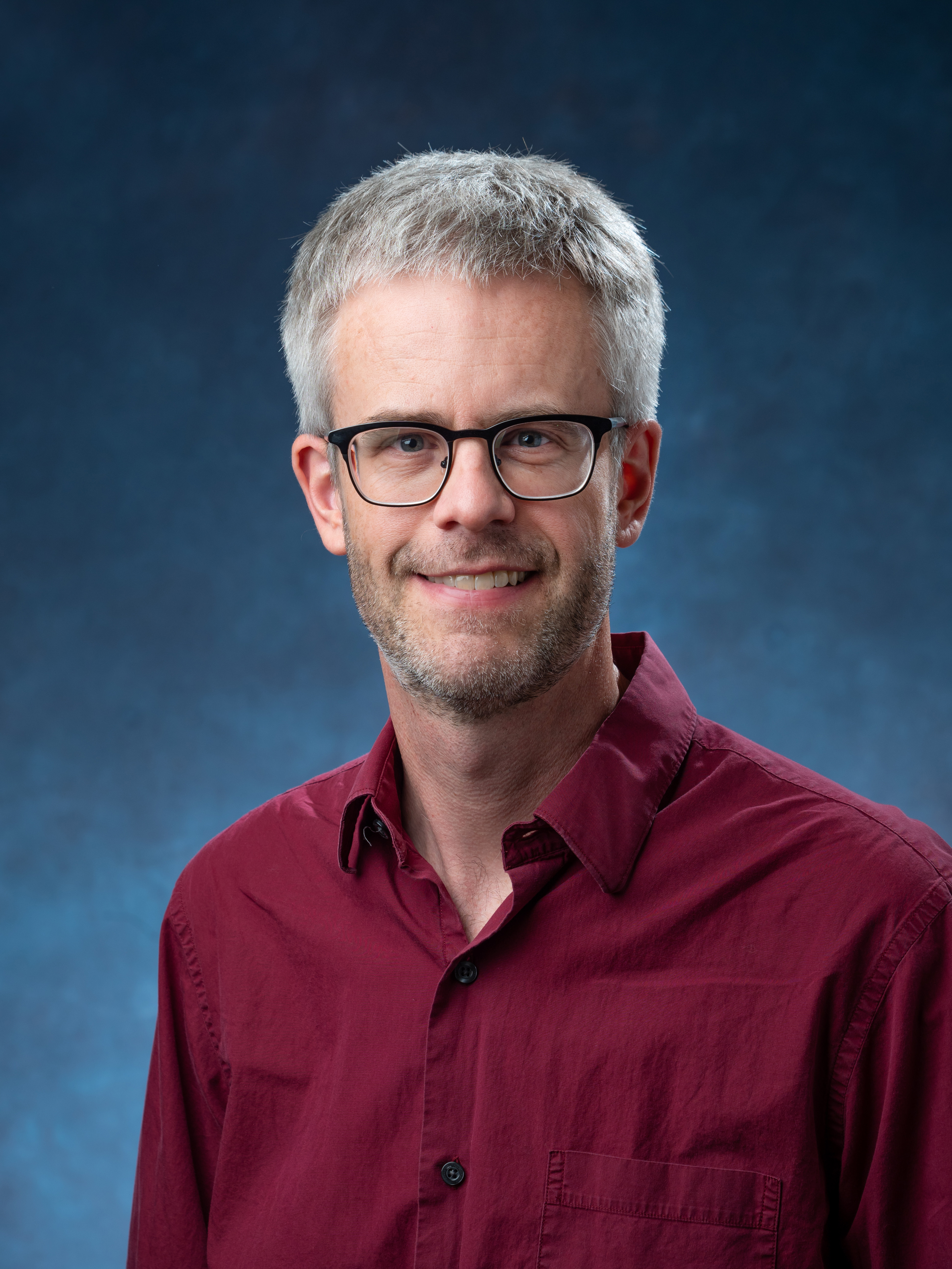 Portrait photo of Michael McGehee smiling at the camera, wearing glasses and a red shirt, against a solid blue background. 