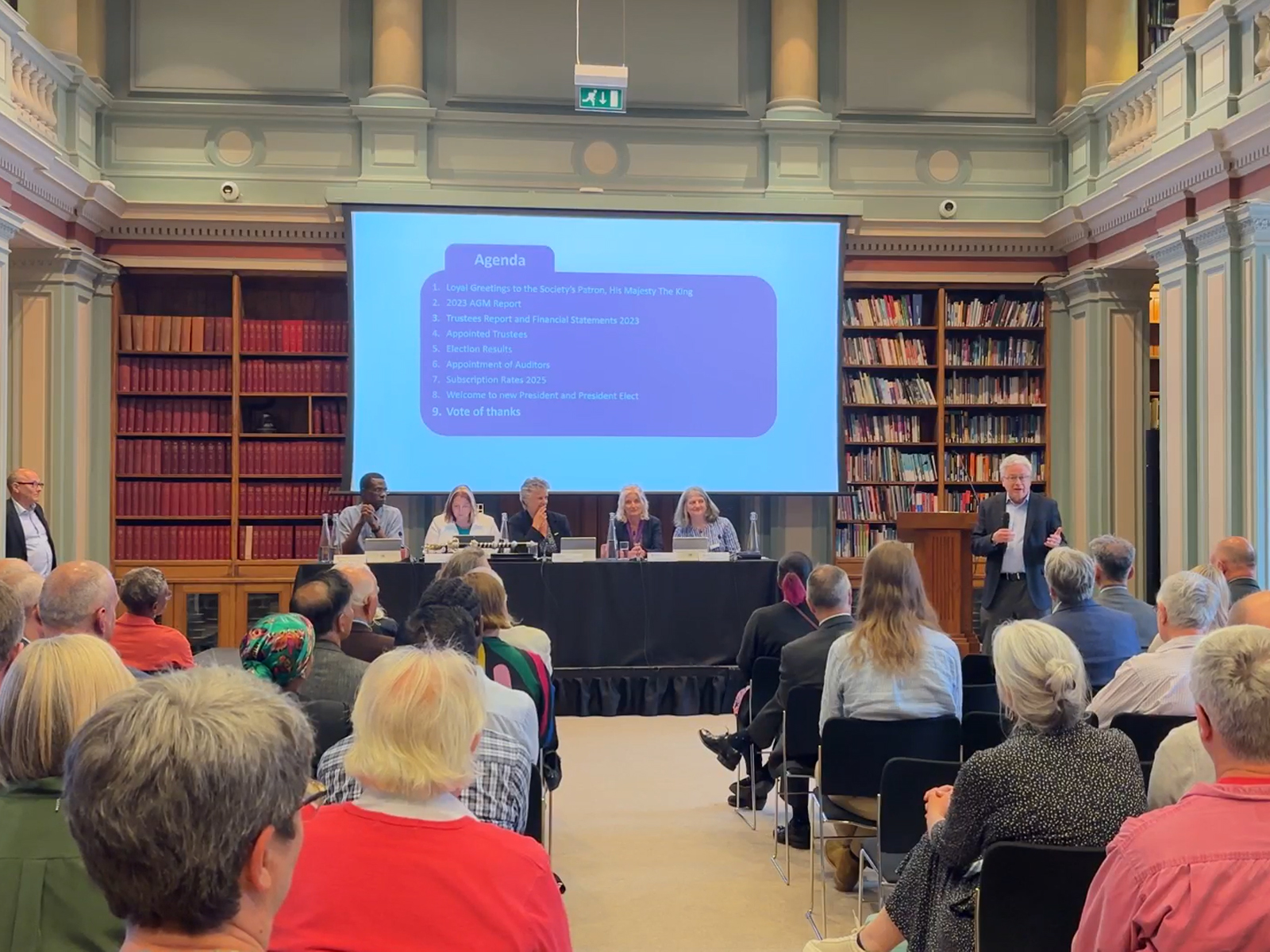 The library at Burlington House, filled with people seated in an audience and a panel of speakers seated on a rostrum at the front of the room