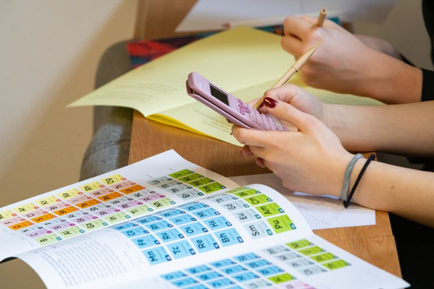 A student holds and works on a calculator while the periodic table lies on a table