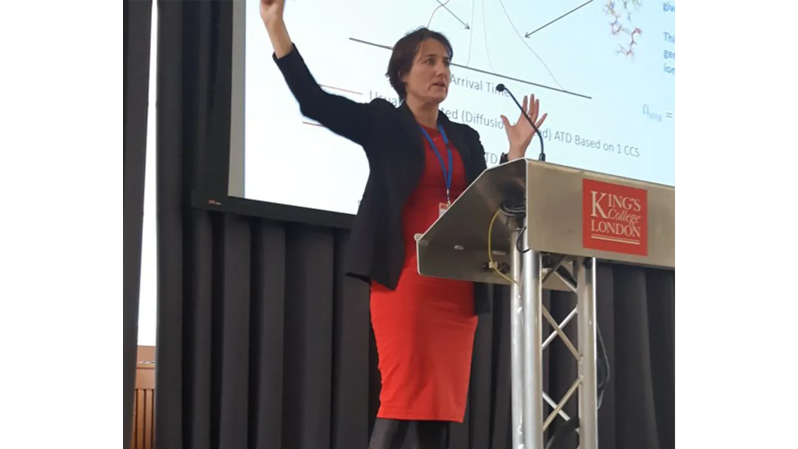 Professor Perdita Barran standing at a podium with her hand up, in front of a screen