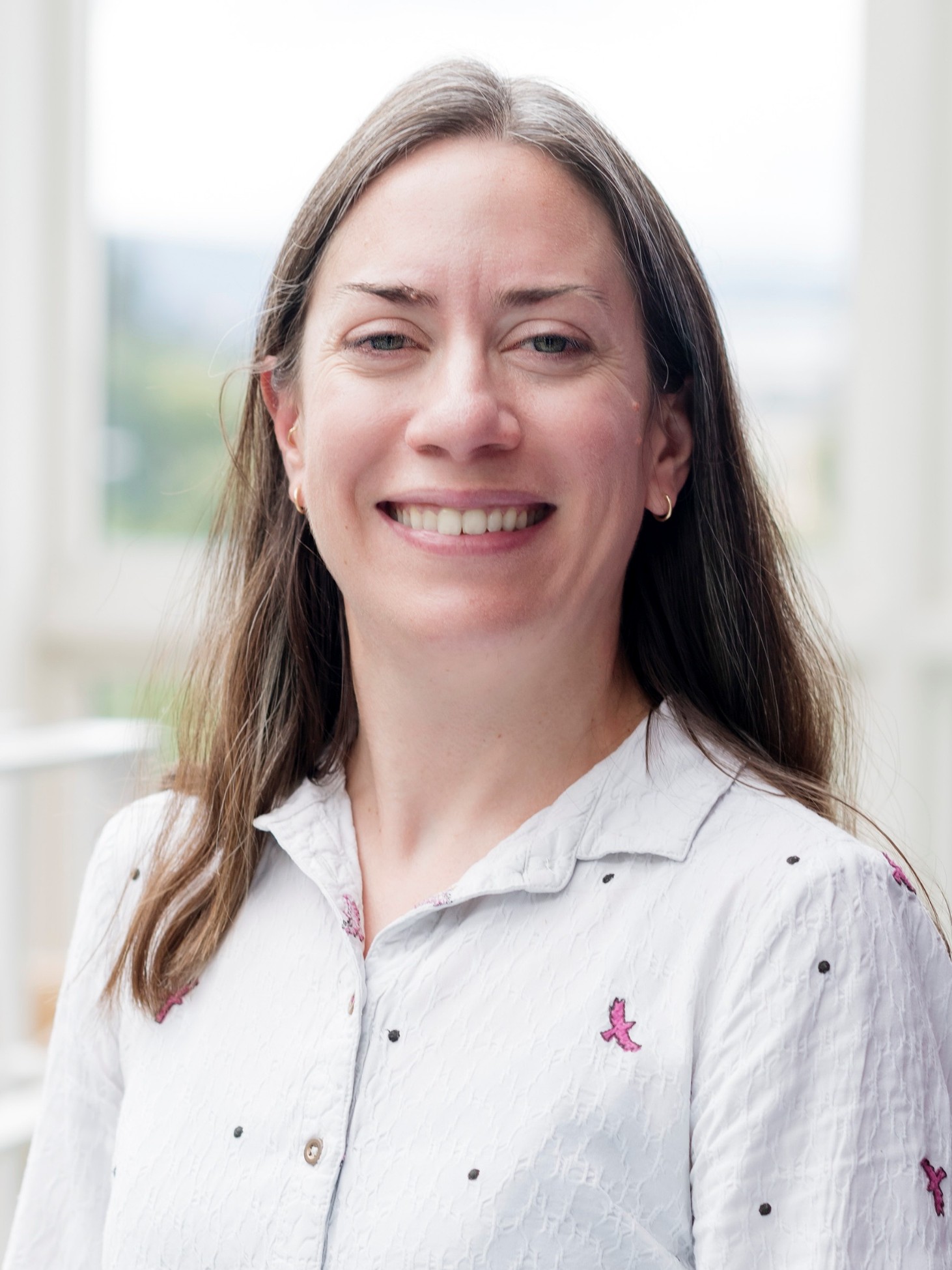 Veronica Sofianos smiling at the camera, wearing a white shirt adorned with small embroidery designs against softly blurred background.